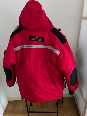 Röd dunjacka Canada Goose Snow Mantra - Röd dunjacka från Canada Goose, modell Snow Mantra. Jackan har stor huva med avtagbar pälskant, flera fickor, reflexdetaljer och svarta förstärkningar på armbågar och axlar. Perfekt för kalla vinterdagar och extrem kyla.