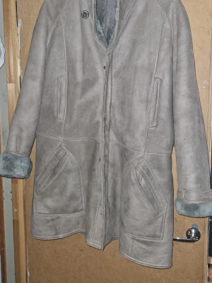 Grå äkta fårskinnskappa med uppvikta ärmar - Snygg grå fårskinnskappa med mjukt foder och uppvikta ärmslut. Kappan har två stora fickor framtill och stängs med knappar. Perfekt för kalla dagar och ger en klassisk look med sin raka passform och diskreta sömmar.