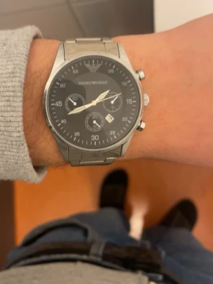 Emporio Armani silverfärgad klocka - Stilren klocka från Emporio Armani med svart urtavla och silverfärgat armband i metall. Klockan har kronograf-funktion, datumvisning och flera små urtavlor. Klassisk design som passar till många olika stilar.