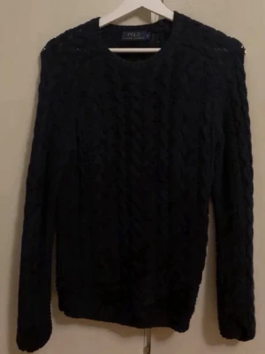 Mörkblå kabelstickad tröja Polo Ralph Lauren - Mörkblå kabelstickad tröja från Polo Ralph Lauren med rund halsringning och långa ärmar. Klassisk och tidlös design med grovstickat mönster, perfekt för lager-på-lager-stil. Snygg att matcha med jeans eller kjol.