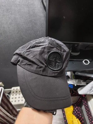 Svart keps från Stone Island - Säljer en svart keps från Stone Island med det klassiska kompassmärket broderat framtill. Kepsen har böjd skärm och är tillverkad i ett lätt, glansigt material som ger en modern look. Perfekt accessoar för dig som gillar streetwear och stilrena detaljer.