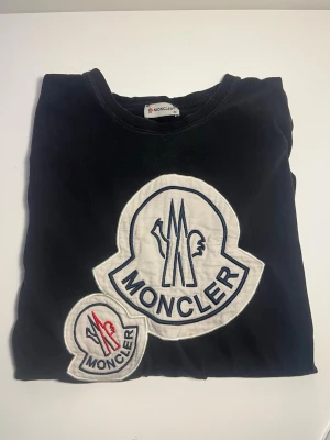 Svart Moncler t-shirt med stora loggor - Säljer en svart t-shirt från Moncler med två stora broderade loggor framtill, en i vitt och blått och en mindre med röd detalj. T-shirten har klassisk rund hals och är i mjuk bomull. Perfekt för dig som gillar statement-plagg och exklusiva märken. Skick 8/10! Skicka gärna bud vill bara få bort den stolek M men passar s