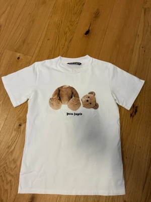 Vit Palm Angels t-shirt med björntryck - Säljer en vit t-shirt från Palm Angels med ett ikoniskt björntryck på framsidan och stor logga i svart på ryggen. Klassisk passform och rund hals. Perfekt för dig som gillar streetwear och vill sticka ut med en unik design.
