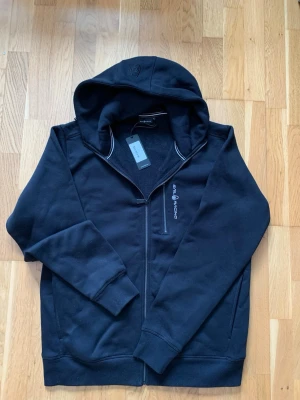 Svart zip hoodie från Sail Racing - Svart hoodie från Sail Racing med dragkedja framtill och broderad logga på bröstet. Tröjan har huva, ribbade muddar och en ficka med dragkedja. Tillverkad i mjukt bomullsmaterial som känns skönt mot huden. Perfekt för en avslappnad och sportig stil. Tröjan är helt oanvänd. Pris kan diskuteras. Tveka inte på att höra av er vid eventuella frågor!
