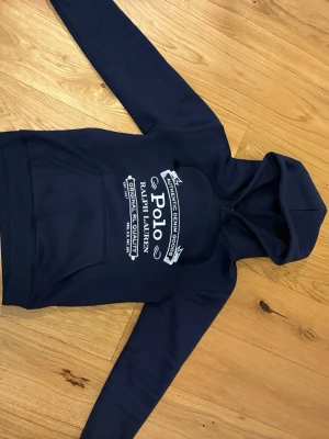 Marinblå hoodie från Polo Ralph Lauren - Snygg marinblå hoodie från Polo Ralph Lauren med vit logga och text på bröstet. Klassisk huva med dragsko och stor magficka framtill. Tillverkad i mjukt material som känns skönt mot huden. Perfekt för en avslappnad och stilren look.