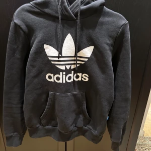 Svart Adidas hoodie med stor logga - Svart hoodie från Adidas med stor vit Trefoil-logga och text på bröstet. Klassisk känguruficka framtill, dragsko i huvan och ribbade muddar. Perfekt för chill eller streetstyle. Mjuk insida och skön passform.