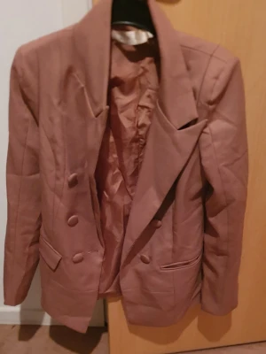 Kavaj - Snygg brun kavaj med dubbelknäppning och breda slag. Kavajen har två framfickor med lock och matchande tygklädda knappar. Tillverkad i ett slätt tyg med helfodrad insida. Perfekt för en trendig och stilren look.