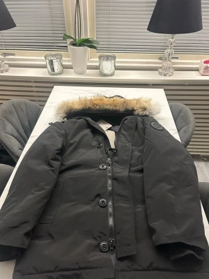 Svart parkas från Canada Goose - Säljer en svart Canada Goose chateau parka storlek M men passar en L med klassisk patch på ärmen och fuskpäls på huvan S. Jackan har dragkedja och knappar framtill samt flera fickor. Perfekt för kalla dagar och riktigt snygg streetstil.