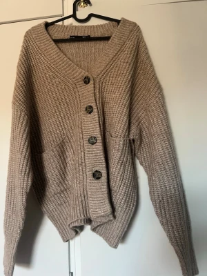 Beige stickad kofta från 157 - Mysig beige stickad kofta från 157 med stora knappar framtill och två fickor. Koftan har en djup v-ringning, ribbad struktur och lång ärm. Perfekt för lager på lager och chill vardagsstil.