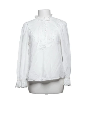 Vit blus med spetsdetaljer - Söt vit blus med volangkrage och spetsdetaljer längs bröstet och ärmsluten. Blusen har lång ärm och en lös passform, perfekt för en trendig och feminin look. Perfekt att ha under en stickad tröja!