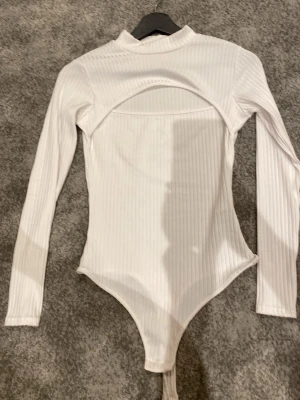 Vit ribbad body från NLY One - Säljer en vit ribbad body från NLY One med lång ärm och hög hals. Bodyn har en snygg cut out-detalj framtill och är tillverkad i stretchigt material som sitter tajt mot kroppen. Perfekt för att skapa en trendig look.