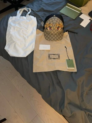 Gucci keps med tigertryck - Snygg keps från Gucci med klassiskt GG-mönster i beige och brunt samt två broderade tigrar framtill. Kepsen har svart mesh på sidorna och justerbar rem bak. Materialet är en mix av canvas och mesh, perfekt för en trendig streetstyle-look.