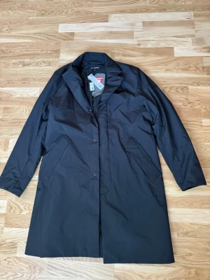 Svart rock från J.Lindeberg - Stilren svart rock från J.Lindeberg med rak passform och dold knappslå. Tillverkad i ett vattenavvisande material med Primaloft-foder som håller dig varm. Klassisk krage och två snedställda fickor framtill. Perfekt för dig som vill ha en modern och enkel look. Nypris ca 4500-5500kr