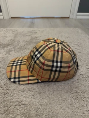 Beige rutig keps från Burberry - Säljer en klassisk beige keps från Burberry med det ikoniska rutmönstret i svart, vitt och rött. Det är den gamla modellen som inte säljs mer så väldigt svår att få tag på idag om inte man köper begagnad 