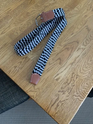 Svartvitt zigzag-mönstrat bälte - Snyggt svartvitt bälte med zigzag-mönster och detaljer i brunt skinn. Bältet har ett klassiskt spänne i silverfärg och passar perfekt för att ge din outfit en cool touch. Materialet är elastiskt och bekvämt.