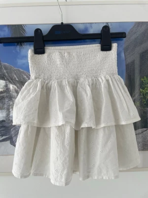 Vit volangkjol med broderade detaljer - Supersöt vit kortkjol med dubbla volanger och broderade hålmönster. Kjolen har en bred, smockad midja som ger en bekväm och snygg passform. Perfekt för sommarens alla tillfällen och lätt att matcha med olika toppar.