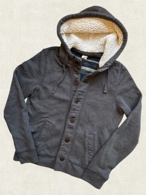 Hollister Cardigan med Luva - En unik cardigan av det populära märket ”Hollister” med en fodrad luva! Extremt bra passform, hör av dig vid frågor.