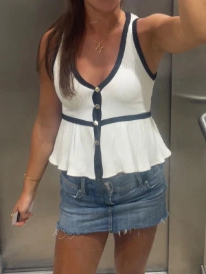 Vit volangtopp med svarta detaljer - Superfin vit ribbad topp med svarta kanter och guldknappar framtill. Toppen har v-ringning, markerad midja och volangig nederdel. Från Zara, går inte längre att köpa. 