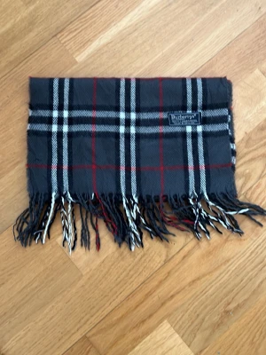 Rutig halsduk från Burberry - Snygg halsduk från Burberry i klassiskt rutigt mönster med svart, vitt och röda linjer. Halsduken har fransar i ändarna och en vävd Burberry-logga. Perfekt accessoar för att lyfta din outfit.