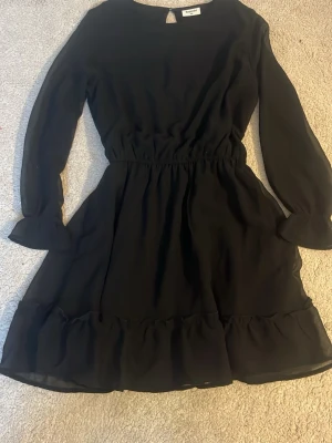 Svart volangklänning med lång ärm - Snygg svart klänning med volangkant nedtill och långa ärmar. Klänningen har en markerad midja och är tillverkad i ett lätt, flödande material. Perfekt för dig som gillar enkel men stilren design. Storlek 158