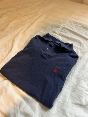 Mörkblå pikétröja från Ralph Lauren - Klassisk mörkblå pikétröja från Ralph Lauren med två vita knappar och röd broderad logga på bröstet. Tröjan har en mjuk krage och är gjord i bomull. Perfekt till jeans eller chinos för en clean och stilren look. Storlek S slim fit
