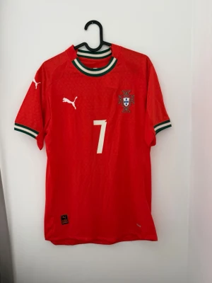 Portugal fotbollströja Ronaldo #7 Puma - Röd Portugal fotbollströja från Puma med nummer 7 och Ronaldo på ryggen. Tröjan har gröna och vita detaljer vid krage och ärmslut samt Portugals landslagsemblem på bröstet. Perfekt för dig som älskar fotboll och vill stötta landslaget.