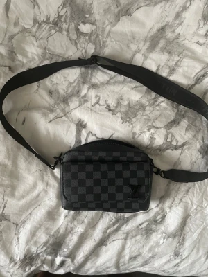 Louis Vuitton axelväska i svart rutmönster - Snygg axelväska från Louis Vuitton i svart och grått med klassiskt rutmönster. Väskan har justerbar axelrem med Louis Vuitton-logga och en kompakt, rektangulär form. Tillverkad i slitstarkt material med diskret LV-detalj på framsidan.