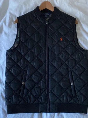 Svart quiltad dunväst från Polo Ralph Lauren - Snygg svart dunväst från Polo Ralph Lauren med quiltat mönster och orange logga på bröstet. Västen har två fickor med tryckknappar och dragkedja framtill. Perfekt för lager på lager och ger en clean look. Insidan camoflauge men syns inte en susning när du bär denna stilrena väst
