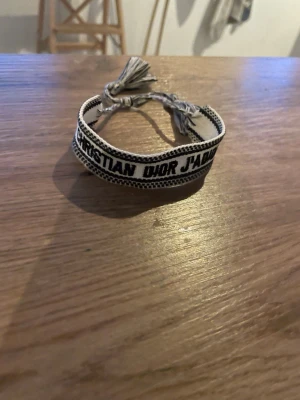 Christian Dior J'adior tygarmband - Snyggt tygarmband från Christian Dior med texten 'J'adior' i svart på beige botten. Armbandet har justerbar knytning med tofsar i ändarna och en cool, bohemisk vibe. Perfekt accessoar för att lyfta vilken outfit som helst.