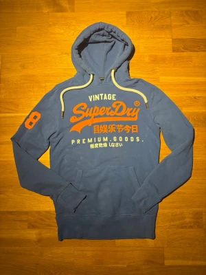 Superdry hoodie - Snygg blå hoodie från Superdry med stor orange logga och japansk text på bröstet. Riktigr snygg och unik. Har inga defekter som man ser på bilderna. Är storlek S men passar även M då den är lite mittemellan. Hör av dig vid frågor om mått eller prisförslag.