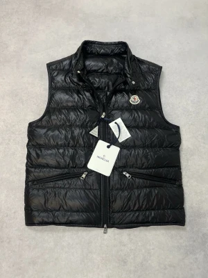 Svart dunväst från Moncler - Snygg svart dunväst från Moncler med klassisk quiltad design och hög krage. Västen har två dragkedjeförsedda fickor framtill och ikonisk Moncler-logga på bröstet. Tillverkad i glansigt material som ger en exklusiv känsla. Perfekt för lager-på-lager-stil.