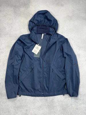 Mörkblå vindjacka med huva från Woolrich - Snygg mörkblå vindjacka med dragkedja framtill och praktisk huva. Jackan har två snedställda fickor och är tillverkad i ett lätt, vattenavvisande material. Perfekt för blåsiga dagar och enkel att matcha med jeans eller andra plagg.