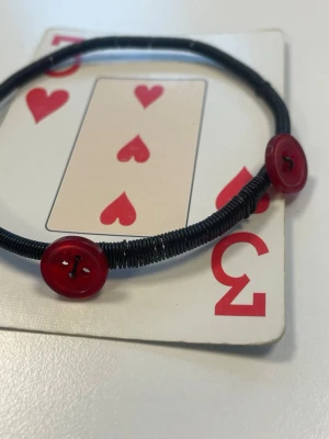 Svart armband med röda knappar - Ett svart armband med två röda knappar som dekoration. Armbandet är gjort av en tunn, svart tråd och har en enkel, minimalistisk design med en lekfull touch tack vare knapparna. Perfekt för dig som gillar unika accessoarer.