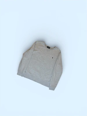 Grå sweatshirt från Polo Ralph Lauren - Klassisk grå sweatshirt från Polo Ralph Lauren med rund halsringning och diskret broderad logga på bröstet. Tröjan har långa ärmar och ribbade muddar vid ärmslut och nederkant. Perfekt basplagg för en avslappnad stil.