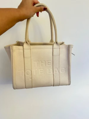 Marc Jacobs Tote Bag - Ej äkta! Marc Jacobs ”The tote bag” i en offwhite/beige färg. Perfekt skick! Långt band där det står Marc Jacobs medföljer. Ett litet innerfack.