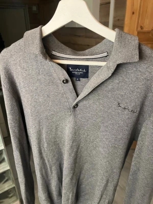 Grå långärmad tröja Bondelid - Snygg grå långärmad tröja från Bondelid med klassisk krage och två knappar vid halsen. Tröjan har diskret broderad logga på bröstet och är tillverkad i mjukt material som känns skönt mot huden. Inget fel på den.