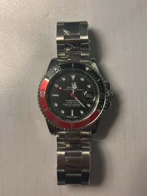  Submariner klocka, rödsvart - Snygg Submariner med svart urtavla, datumfönster och röd-svart vridbar bezel. Klockan har en robust länk i rostfritt  och tydliga indexmarkeringar. Perfekt för dig som gillar klassisk och sportig stil.