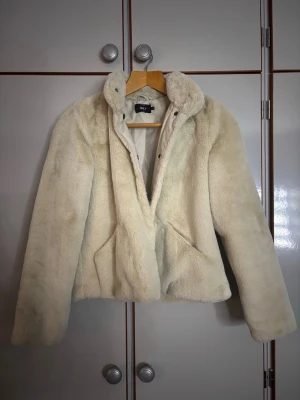Beige fuskpälsjacka från Only - Snygg beige fuskpälsjacka från Only med hög krage och dold knäppning. Jackan har två snedställda fickor framtill och är helfodrad för extra värme. Perfekt för att hålla stilen under kalla vinterdagar.