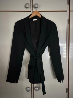 Svart kavaj med knytskärp från Gina Tricot - Stilren svart kavaj från Gina Tricot med lång ärm och markerad midja tack vare ett brett knytskärp. Kavajen har klassisk krage och rak passform, perfekt för en trendig look. Materialet är mjukt och följsamt.