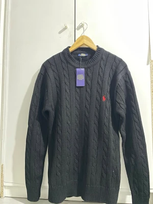 Svart kabelstickad tröja Polo Ralph Lauren - Kabelstickad svart tröja från Polo Ralph Lauren med rund halsringning och röd broderad logga på bröstet. Helt ny