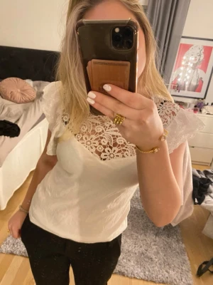 Vit blus med spetsdetaljer - Säljer en söt vit blus med blommig spets upptill och volangärmar. Perfekt att styla med jeans eller kjol för en trendig outfit.