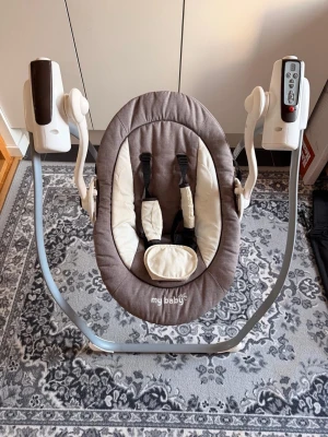 Elektrisk babysitter  - Elektrisk babysitter i grått och vitt från My Baby. Sitsen har mjuk stoppning, säkerhetsbälte och justerbara funktioner via kontrollpanel. Stativet är i metall med plastdetaljer och stolen har ergonomisk form för bekväm vila. Perfekt för att vagga och underhålla små barn 3-6 månader. 