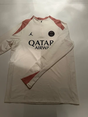Vit PSG träningströja Jordan - Snygg långärmad träningströja från PSG i samarbete med Jordan. Tröjan är vit med rosa detaljer på axlar och ärmslut, halv dragkedja och tryckta loggor på bröstet samt texten PARIS på ärmen. Perfekt för fotbollsträning eller chill.