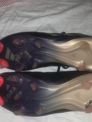 Adidas Copa fotbollsskor svart/röd - Säljer ett par Adidas Copa fotbollsskor i svart med röda detaljer och klassisk design. Skorna har fasta dobbar och ovandel i mjukt läder för bra bollkontroll. Insidan är röd och sulan har Copa-loggan. Perfekta för dig som vill ha stilrena och bekväma skor på planen. Finns en slitning på ena skon(se bild 4) annars inga defekter. Köpta för 2500 och är sparsamt använda.