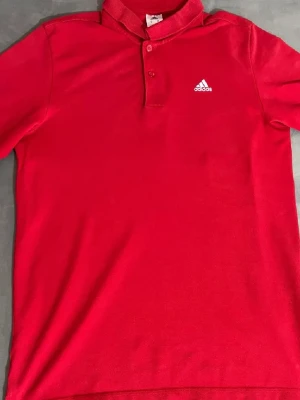 Röd pikétröja från Adidas - Snygg röd pikétröja från Adidas med klassisk krage och knäppning framtill. Tröjan har korta ärmar och är tillverkad i bomullsmix med diskret Adidas-logga broderad på bröstet. Perfekt för en sommar stil.