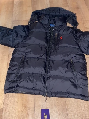 Svart dunjacka från Polo Ralph Lauren - Snygg svart dunjacka från Polo Ralph Lauren med röd logga på bröstet. Jackan har hög krage, dragkedja framtill och två stora fickor med dragkedja. Puffig modell som håller värmen och har en klassisk streetwear-look.