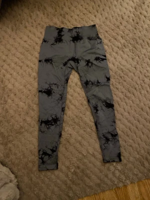 Gym byxor  - Säljer ett par Snygga grå gymbyxor med svart tie-dye mönster, perfekta för träning eller chill. De har hög midja och är tillverkade i ett stretchigt material som sitter skönt på kroppen. Passar dig som vill ha en unik look på gymmet eller till vardags. Byxorna är i storlek L men passar mig som har storlek S i byxor. Har bara använt de 1 gång men sen har de inte kommit till användning. Helt nya utan prislapp. 