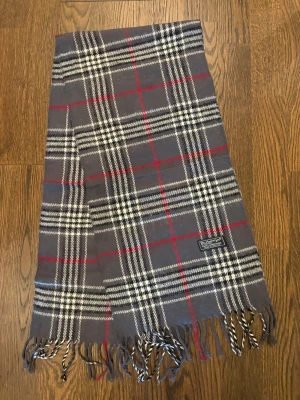Grå Burberry Halsduk - Klassisk halsduk i grått med vitt, svart och rött rutmönster från Burberry. Tillverkad i mjuk ull som känns lyxig och varm. 