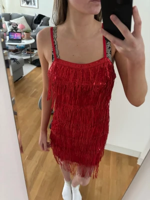 Röd fransklänning med paljetter - Säljer en kort röd klänning med fransar och paljetter. Klänningen har smala axelband med spetsdetaljer och en rak urringning. 