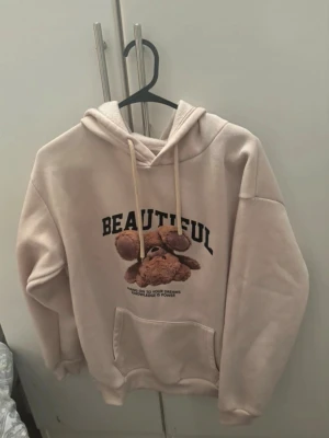 Beige hoodie med nalle och text - Beige hoodie med huva och dragsnören. På bröstet finns en söt nallebjörn och texten 'BEAUTIFUL' i svart samt ett mindre tryck under. Hoodien har en stor magficka och avslappnad passform, perfekt för en chill och trendig look.